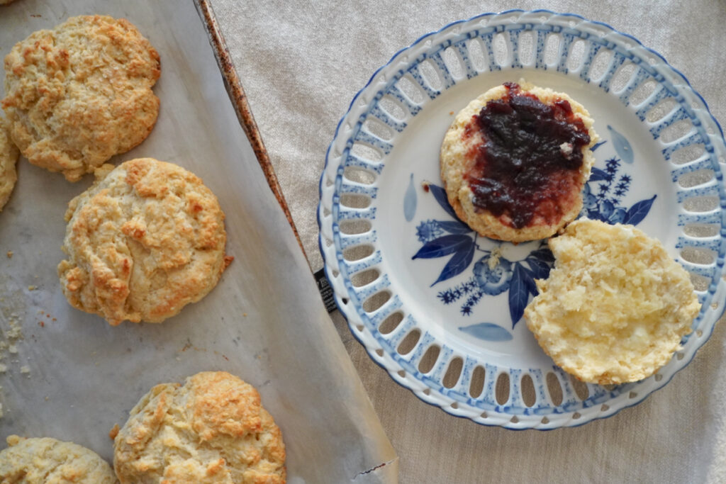 a plate with a buttery biscuit cut in half with butter and blackberry jam next to a baking sheet of more buttery biscuits