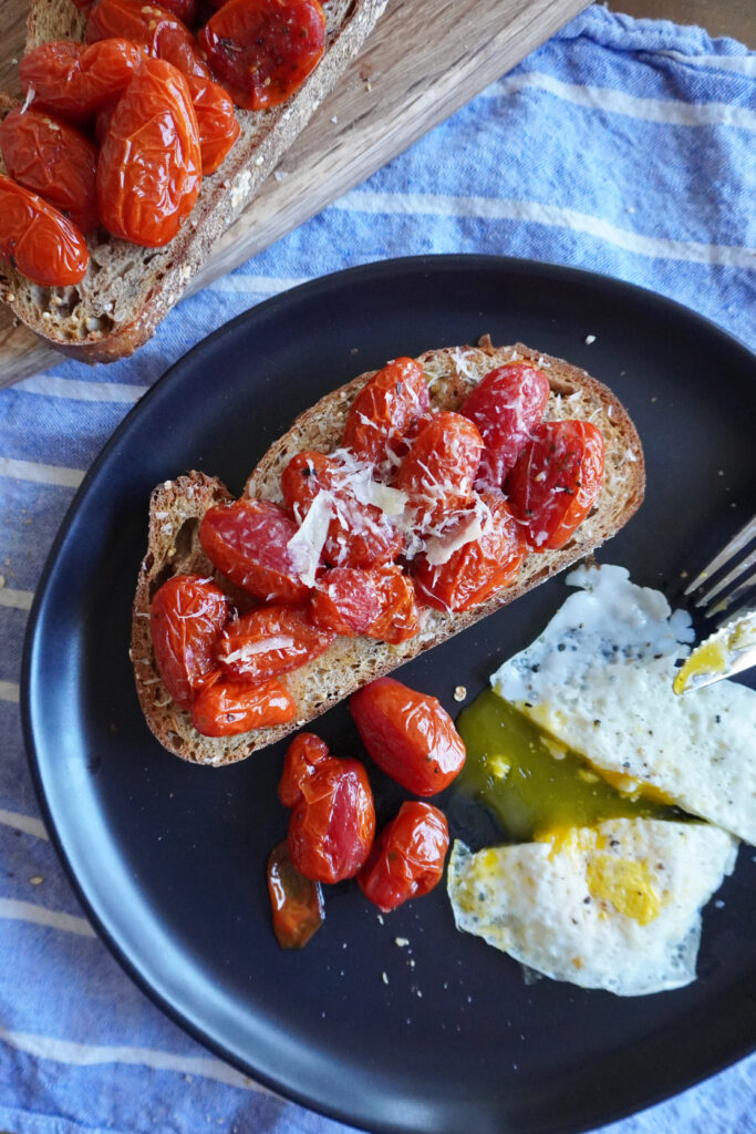 a black plate with roasted grape tomatoes and parmesan cheese on multigrain toast