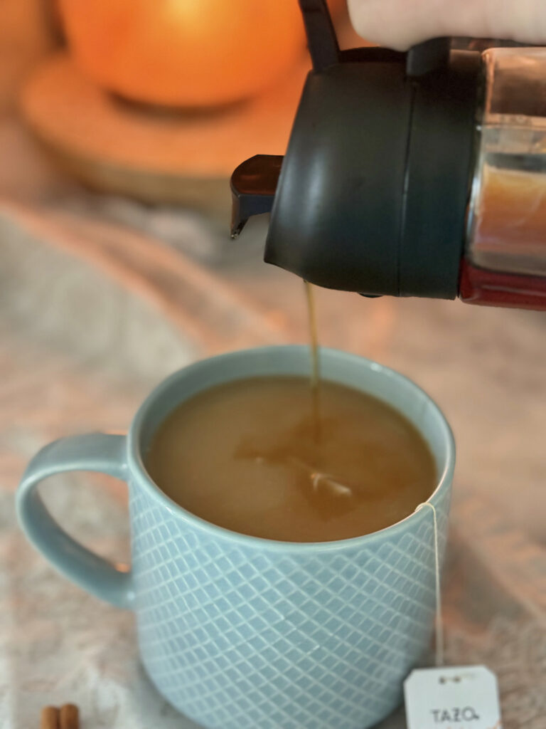 maple syrup being poured from a pitcher into a blue mug of a tea latte