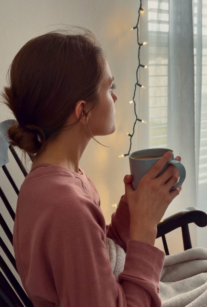 young woman in a pink long-sleeved shirt, with hair in a low bun, holding a blue mug of tea while sitting in a black rocking chair with a blanket