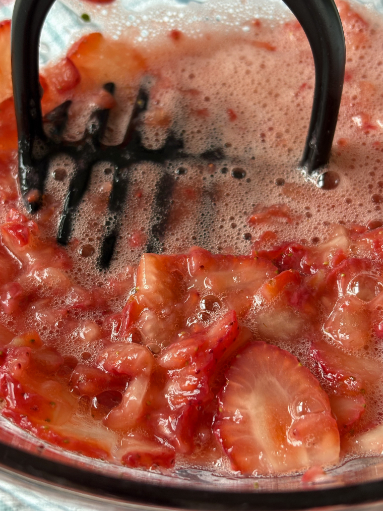a black potato masher mashing strawberries in a glass bowl