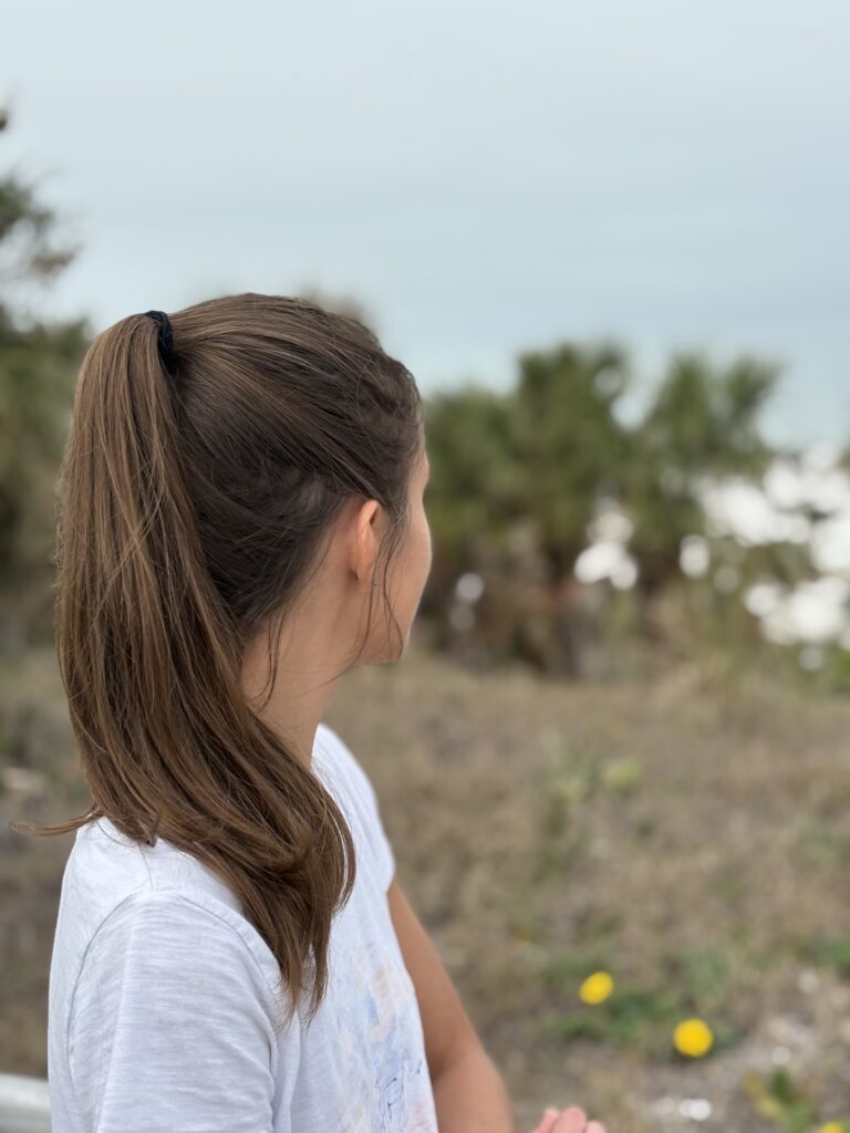woman in white shirt with brown hair in a ponytail looking out at the ocean